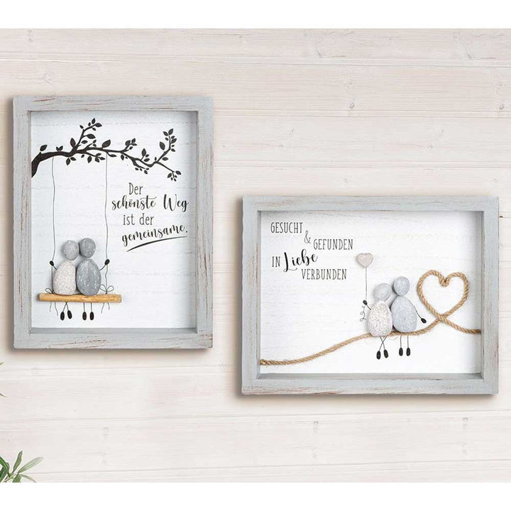 Das Set „Gilde - Steinbild Deko Liebevolles Wandbild mit Spruch“ umfasst zwei gerahmte Wandkunststücke mit Kieselsteinfiguren, eine auf einer Schaukel und eine andere mit einem Herz aus Seil, beide mit deutschem Text auf weißem Holzhintergrund.