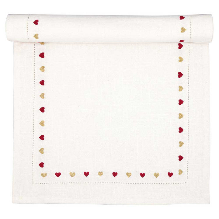 Ein GreenGate - Tischläufer Grace weiß mit Stickerei, verziert mit rot-goldener herzförmiger Stickerei entlang der Kanten, an einem Ende teilweise gerollt.