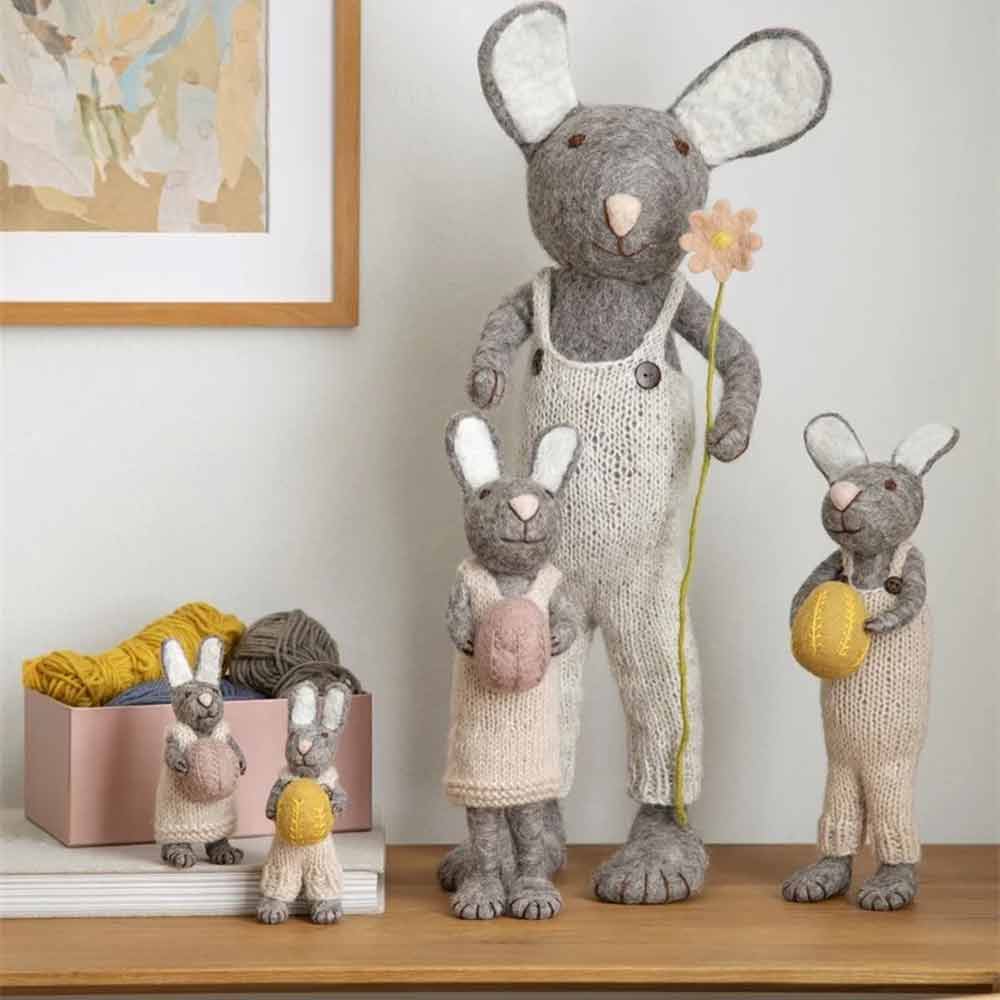 Eine Sammlung hellgrauer Figuren von Gry & Sif – Hase Filz mit Ei, ausgestellt auf einem Regal neben etwas Garn und einem Blumengemälde.