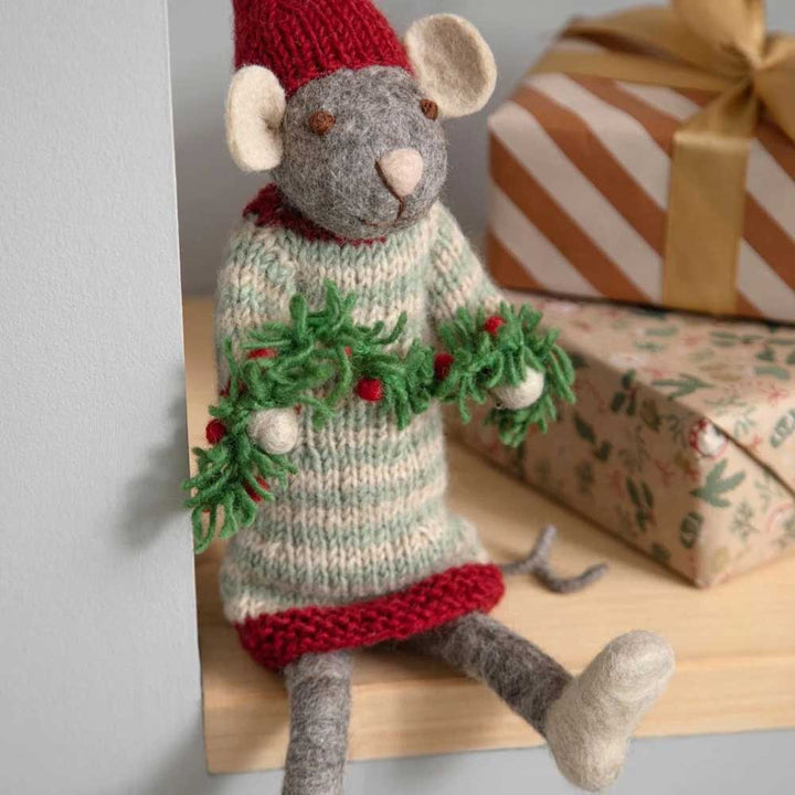 Ein Gry & Sif - Maus Mädchen mit Girlande aus Filz steht auf einem Regal, geschmückt mit einer roten Mütze und einem gestreiften Pullover, und hält eine grüne Girlande. Im Hintergrund sind in braunes Papier eingewickelte Geschenkboxen zu sehen.