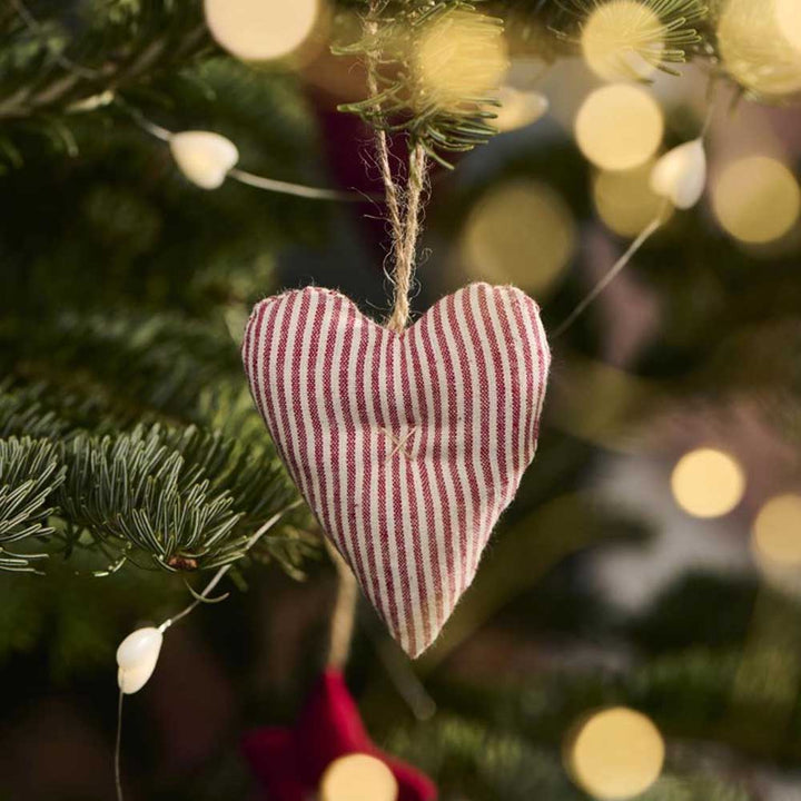 An Ib Laursen - Anhänger Herz Stoff. Mein nostalgisches Weihnachtsornament in roten und weißen Streifen hängt an einem Weihnachtsbaum mit verschwommenen Lichtern im Hintergrund.