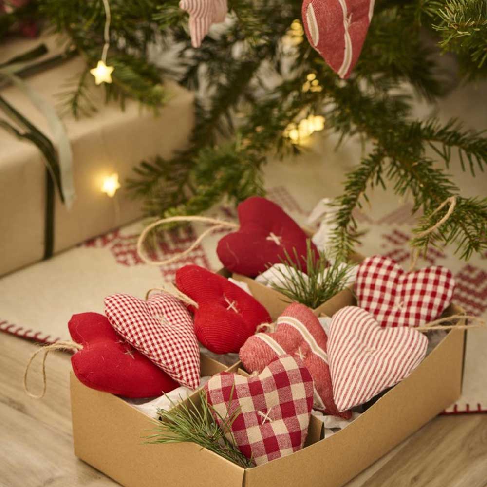 Eine Schachtel mit nostalgischen Weihnachtsdekorationen von Ib Laursen – Anhänger Herz Stoff. In Form von roten und karierten Herzen, platziert unter einem dekorierten, mit Lichtern geschmückten Weihnachtsbaum.