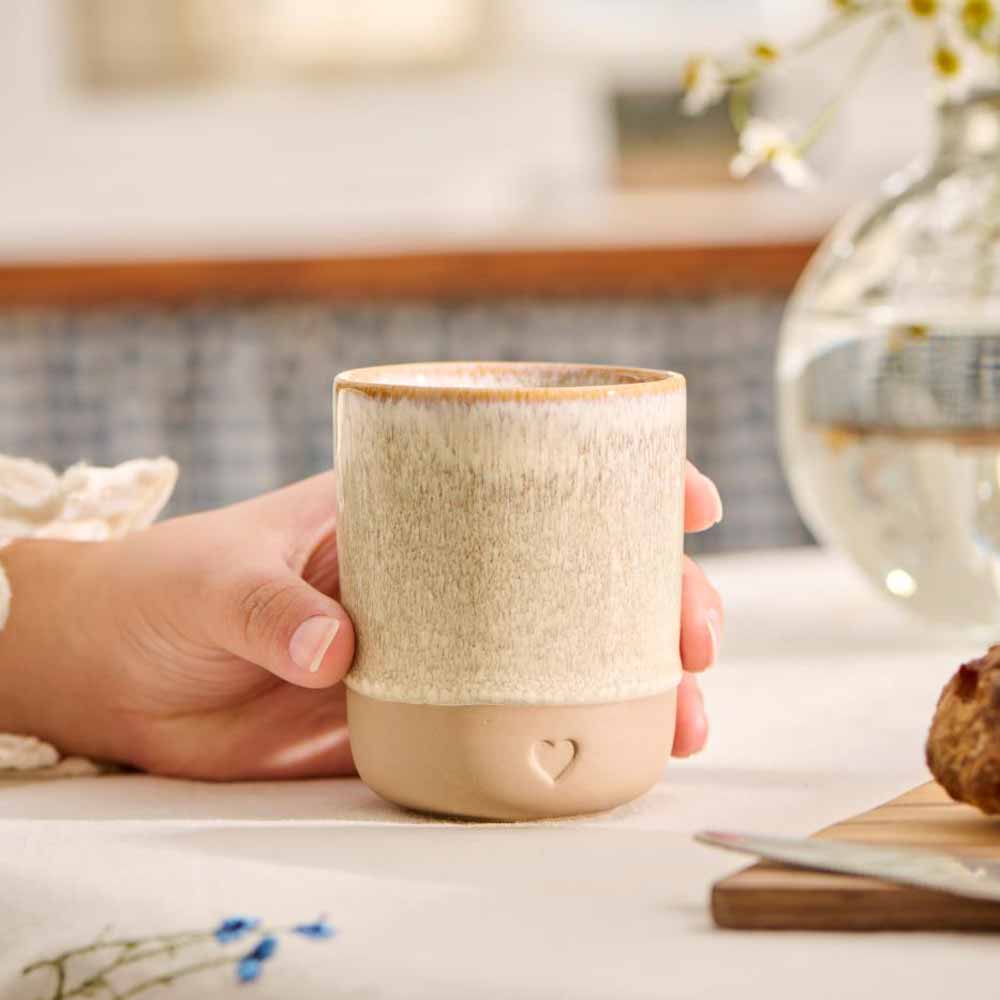 Eine Hand hält einen Ib Laursen - Becher Herz Ton Natur, einen beigefarbenen Keramikbecher mit einem kleinen Herzmotiv am Boden. Im Hintergrund ist ein unscharfer Tisch mit einer Glasvase und Blumen zu sehen.