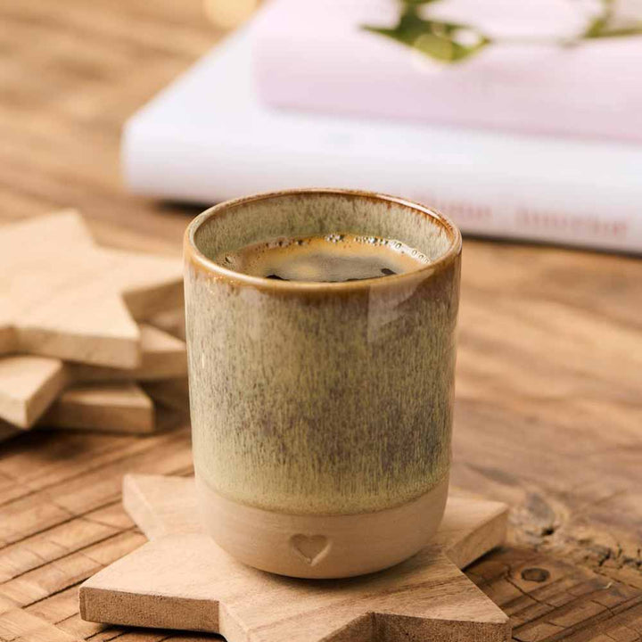 Der Ib Laursen - Becher Herz Ton Natur steht mit Kaffee auf einem hölzernen sternförmigen Untersetzer auf einem Tisch, im Hintergrund sind verschwommene Bücher zu sehen.