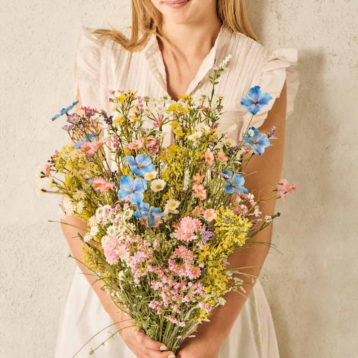 Eine Frau in einem weißen Kleid hält den Blumenstrauß „Blume rosa/grün“ von Ib Laursen mit verschiedenen Wildblumen, darunter rosa und grüne Blüten, vor einer hellen Wand.