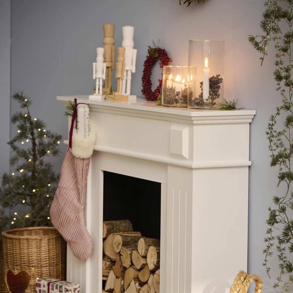 Ein weißer Kaminsims, dekoriert mit Kerzen in Glashaltern, Ib Laursens „Figur Nussknacker abgerundete Füße weiß“ und einem roten Strumpf. Darunter sind im Kamin Holzscheite gestapelt und daneben ein Korb mit verpackten Geschenken.