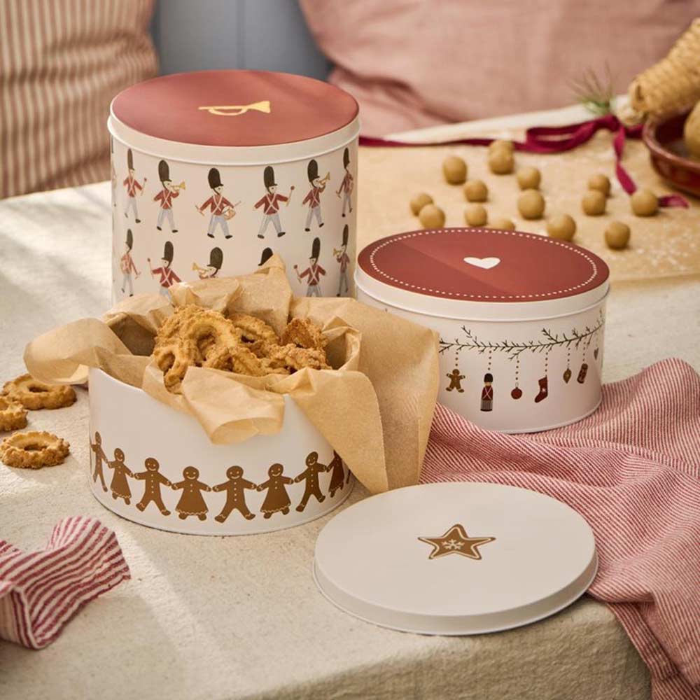 Auf einem Tisch stehen drei runde Dosen mit der Aufschrift „Ib Laursen - Gebäckdose mit Girlande My Nostalgic Christmas“. Jede Dose ist mit Soldaten, Herzen und Lebkuchenfiguren bedruckt. Im Hintergrund sind Plätzchenzutaten zu sehen.