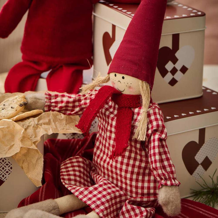 Neben der mit gewebten Weihnachtsherzen verzierten Keksdose „Ib Laursen – Gebäckdose mit geflochtenen Weihnachtsherzen“ sitzt eine Stoffpuppe mit rotem Hut und kariertem Kleid.