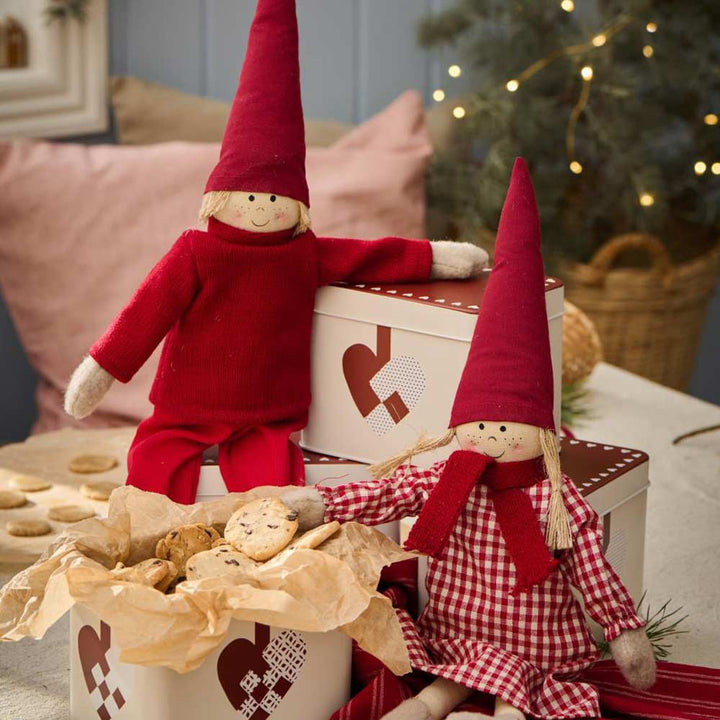 Zwei Plüschfiguren in roten Outfits sitzen auf dekorierten Kisten, umgeben von einer Ib Laursen - Gebäckdose mit geflochtenen Weihnachtsherzen und einem beleuchteten Weihnachtsbaum.