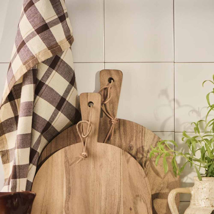 Zwei hölzerne Schneidebretter und ein Ib Laursen - Geschirrtuch Malthe natur mit dunkelbraunem Streifenmuster lehnen an einer gefliesten Wand, auf der rechten Seite ist eine grüne Pflanze teilweise sichtbar.