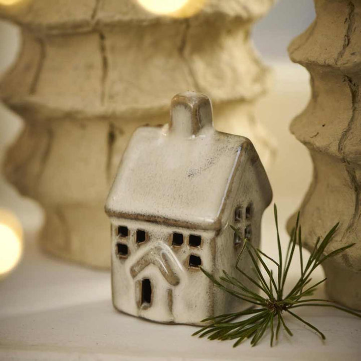 Das Ib Laursen - Haus Mini Vintage Keramikornament ist zwischen zwei verschwommenen Dekobäumen platziert, davor steht ein Tannenzweig.