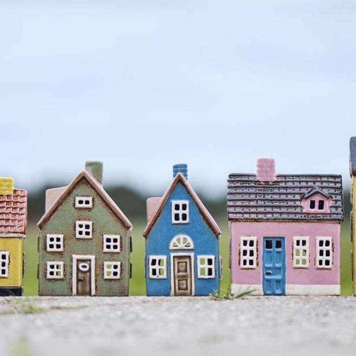 Fünf Ib Laursen „Haus für Teelicht My Home Town blaue Tür“-Kerzenhalter stehen im Freien auf einer ebenen Fläche, im Hintergrund sind ein unscharfer Himmel und ein Feld zu sehen.
