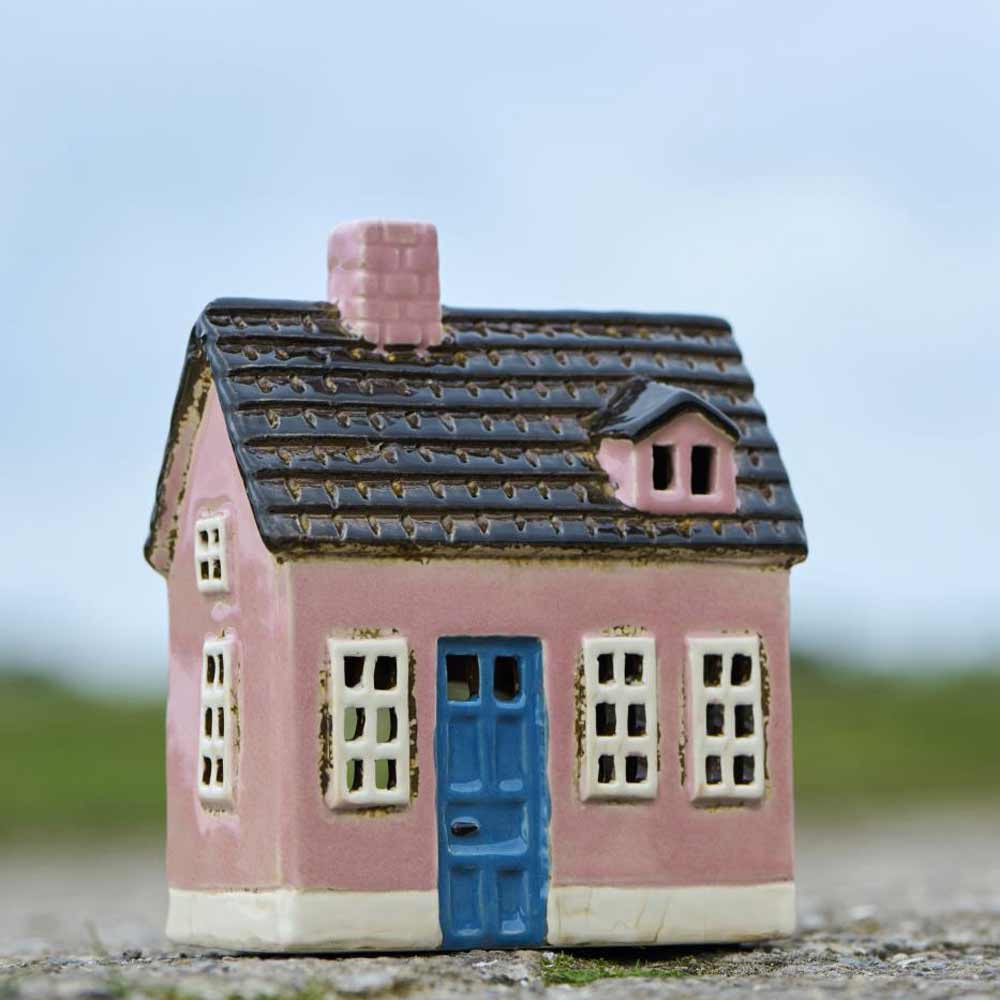 Das Ib Laursen Haus für Teelicht „My Home Town blaue Tür“ ist ein kleines rosa Keramikhaus mit einer blauen Tür und weiß umrandeten Fenstern, das im Freien vor einem unscharfen Hintergrund ausgestellt ist.