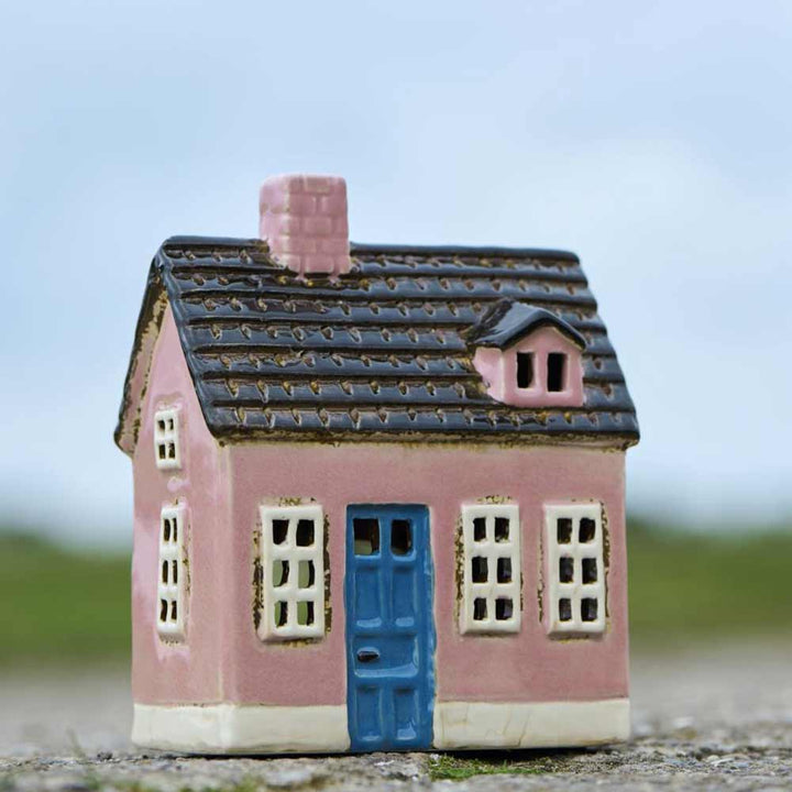 Das Ib Laursen Haus für Teelicht „My Home Town blaue Tür“ ist ein kleines rosa Keramikhaus mit einer blauen Tür und weiß umrandeten Fenstern, das im Freien vor einem unscharfen Hintergrund ausgestellt ist.