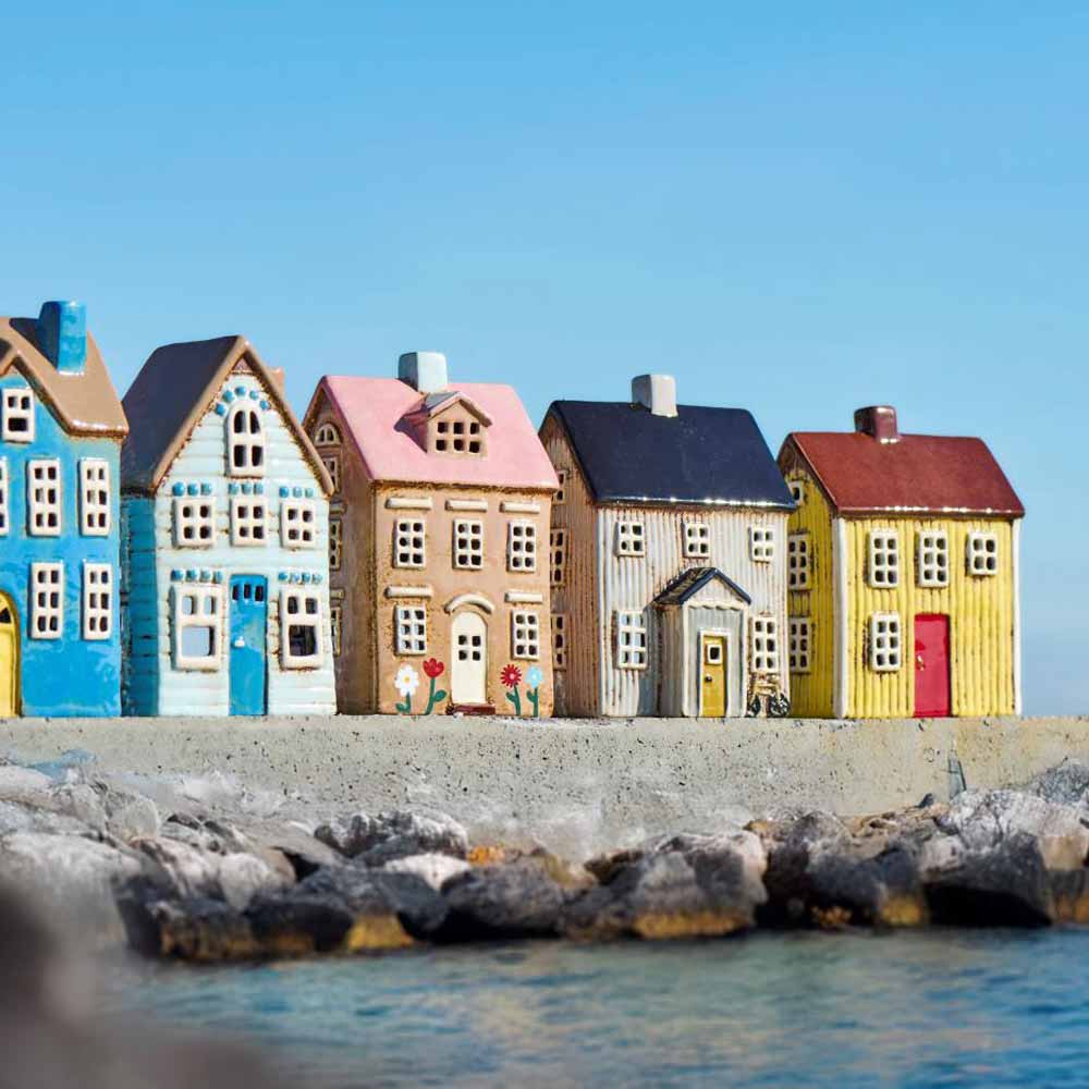 Das Ib Laursen Haus für Teelicht Nyhavn Blumen mit rosa Dach steht auf einer Betonplattform am Wasser, im Vordergrund liegen Steine und im Hintergrund ist ein klarer Himmel zu sehen.