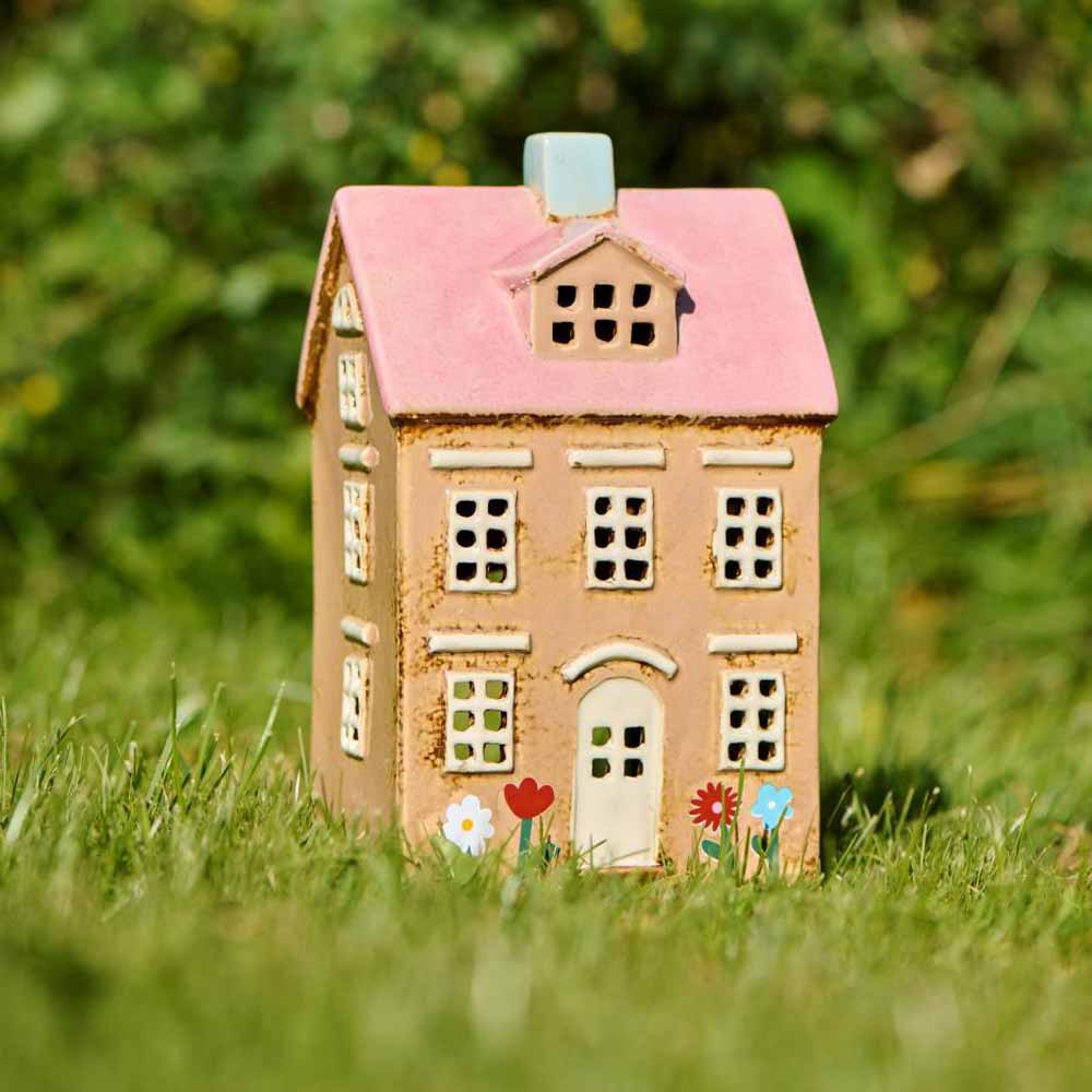 Das Ib Laursen Teelichthaus Nyhavn hat ein rosa Dach, steht auf Gras mit Blumen an der Basis und wird von grünem Hintergrund umgeben.