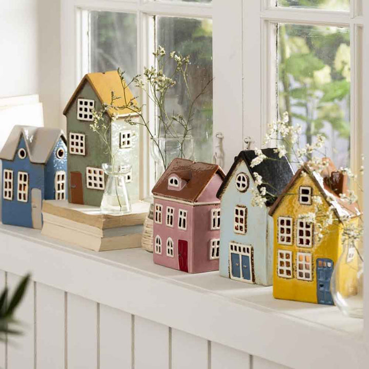Kleine Ib Laursen - Haus für Teelicht Nyhavn gerundete Dachfenster mit unterschiedlichen Farben und Designs stehen auf einem sonnenbeschienenen Fensterbrett neben einem Stapel Bücher und Glasvasen mit zarten Blumen.