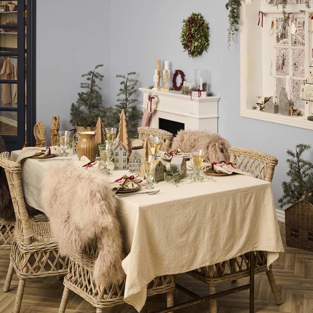 Ein Esstisch mit Geschirr und Weihnachtsdekoration, umgeben von Korbstühlen, die mit Felldecken geschmückt sind. Der Raum hat ein festliches Ambiente mit dem Ib Laursen - Haus für Teelicht Stillenat, das zwischen den verschiedenen Weihnachtsdekorationen und einem Kamin im Hintergrund ein warmes Licht erzeugt.