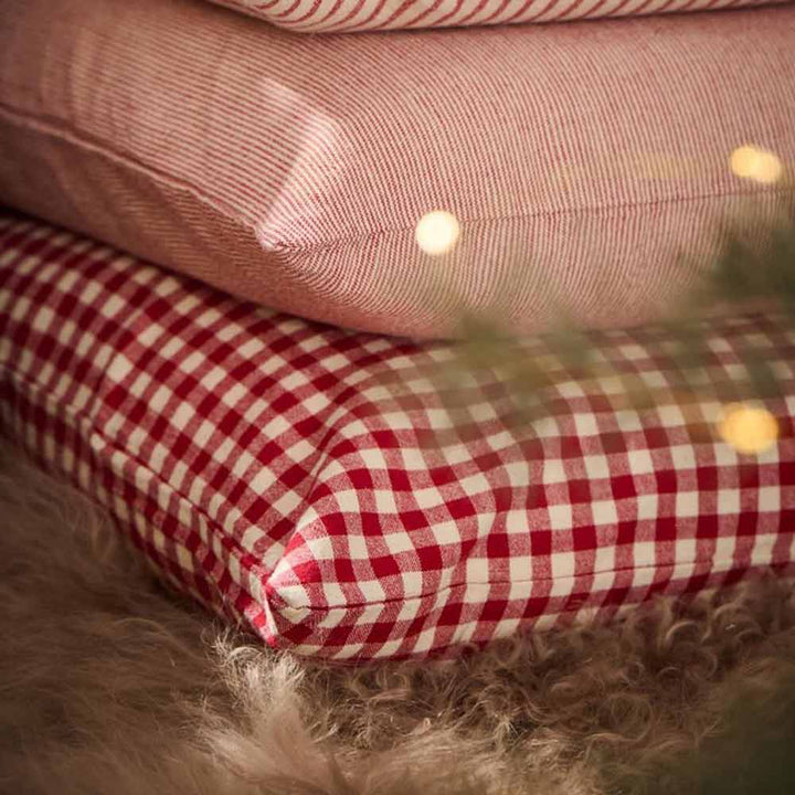 Drei gestapelte Ib Laursen Asta Kissenbezüge in Rot mit kleinen naturfarbenen Karomustern, platziert auf einer flauschigen Unterlage. Verschwommene Lichter und teilweise Grün im Vordergrund sichtbar.