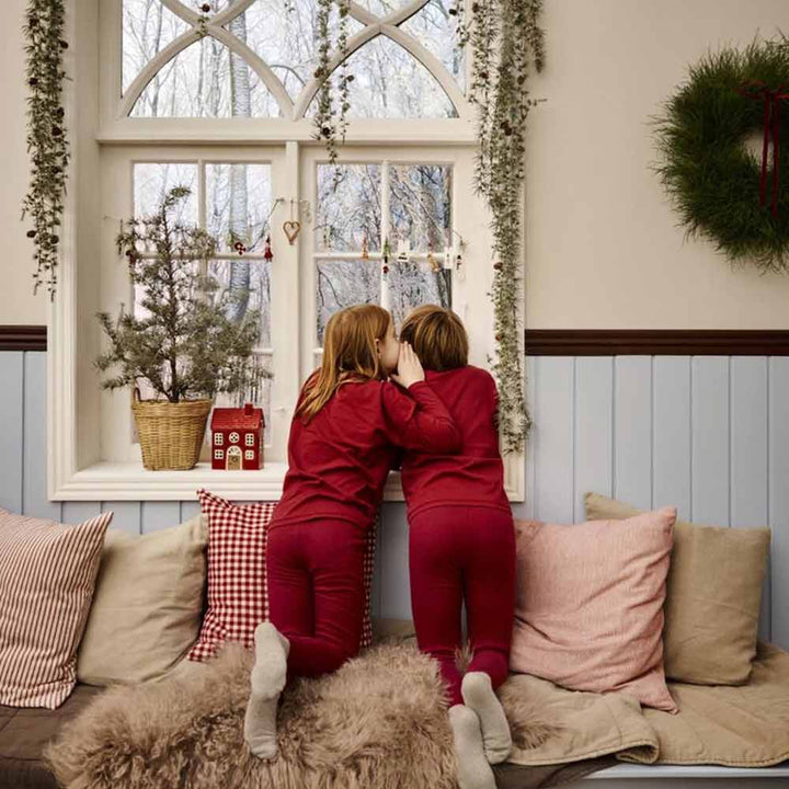 Zwei Kinder in passenden roten Pyjamas lehnen auf einem gepolsterten Fenstersitz mit dem Ib Laursen - Kissenbezug Asta rot mit kleinen naturfarbigen Karos und blicken nach draußen durch ein dekoriertes Fenster mit Topfpflanzen und Weihnachtsschmuck.