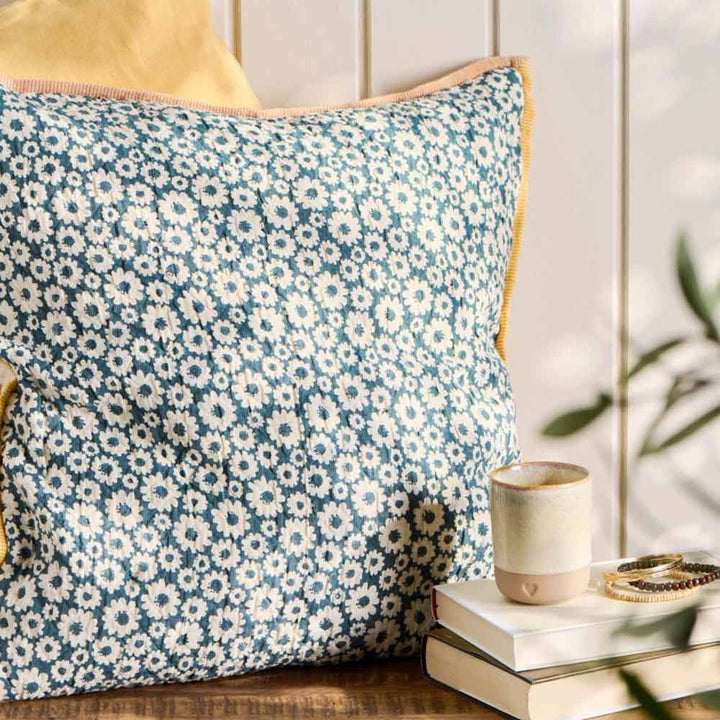 Ein Ib Laursen „Kissenbezug Vigga mit Blümchen Rückseite neutral“ schmückt eine Holzbank und gibt teilweise den Blick auf ein gelbes Kissen frei. Daneben liegen gestapelte Bücher, eine cremefarbene Tasse und ein Armband.