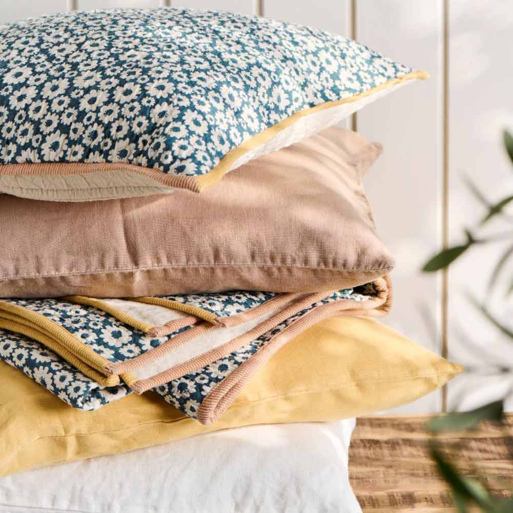Ein Stapel aus vier Ib Laursen Vigga-Kissenbezügen mit floralen und einfarbigen Mustern in Blau, Beige und Gelb ruht auf einer Holzoberfläche.