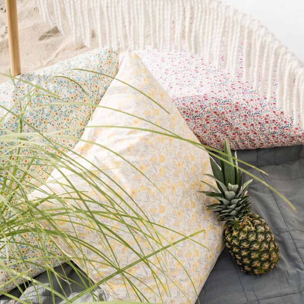 Eine Ananas liegt auf einer grauen Decke neben dem Ib Laursen Kissenbezug in hellrosa und beigem Blumenmuster mit blauen Blättern, gemusterten Kissen, Fransen und im Vordergrund sichtbaren grünen Pflanzenblättern.