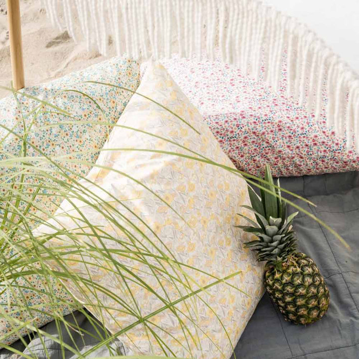 Eine Ananas liegt auf einer grauen Decke neben dem Ib Laursen Kissenbezug in hellrosa und beigem Blumenmuster mit blauen Blättern, gemusterten Kissen, Fransen und im Vordergrund sichtbaren grünen Pflanzenblättern.