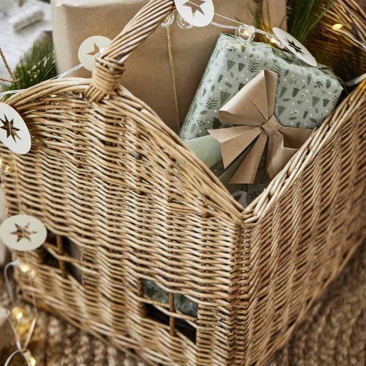 Das Ib Laursen - Korbhaus, ein Weidenkorb gefüllt mit in Papier verpackten Geschenken, darunter ein grünes Päckchen mit beiger Schleife, ist mit Lichterketten und sternförmigen Dekorationen geschmückt.