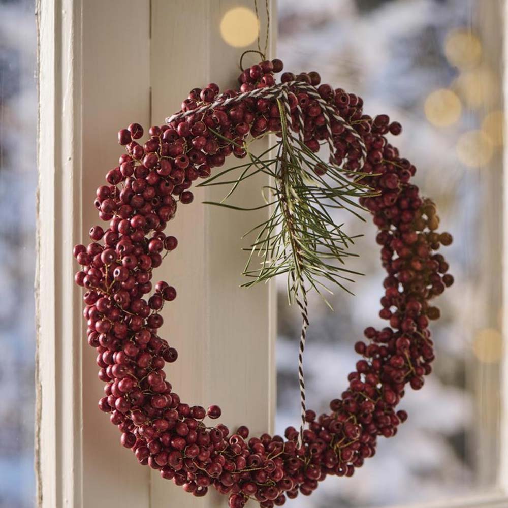 Ein Ib Laursen – Kranz mit Holzperlen Stillenat handgefertigt hängt an einem Fenster und präsentiert seine roten Beeren und Kiefernzweige vor einem sanft verschwommenen, verschneiten Hintergrund.