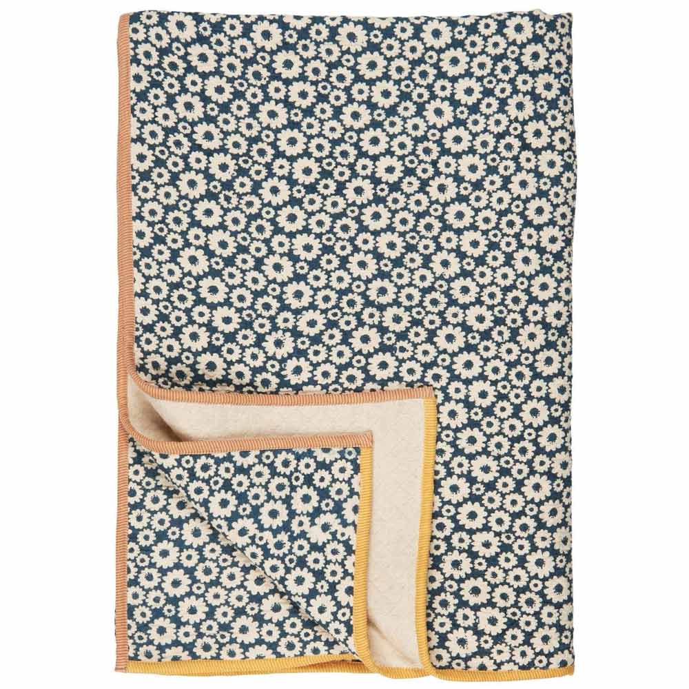 Das Ib Laursen - Plaid Vigga hat ein blau-weißes Blumenmuster mit gänseblümchenartigen Blüten, eine einfarbig beige Unterseite und einen gelb-orangen Rand.