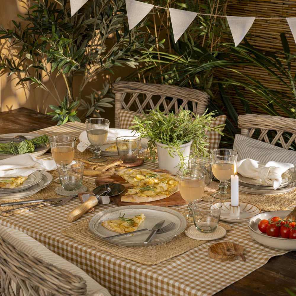 Gedeckter Tisch für eine Mahlzeit im Freien mit Salat-/Pastateller Sand Dunes von Ib Laursen, Besteck, Gläsern und einer Pizza auf einem Holzbrett. Eine Topfpflanze und Kerzen dienen als Dekoration, umgeben von Grünpflanzen und Wimpelketten.