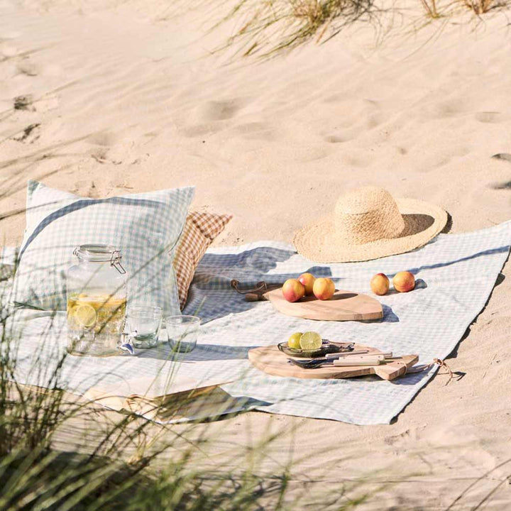 Picknickaufbau am Sandstrand mit karierter Decke, Kissen, Strohhut, Ib Laursen Schneidebrett mit Lederschnur rund Fisch Akazienholz mit Früchten, einem Glas Zitronenwasser und Gläsern. Gras im Vordergrund.