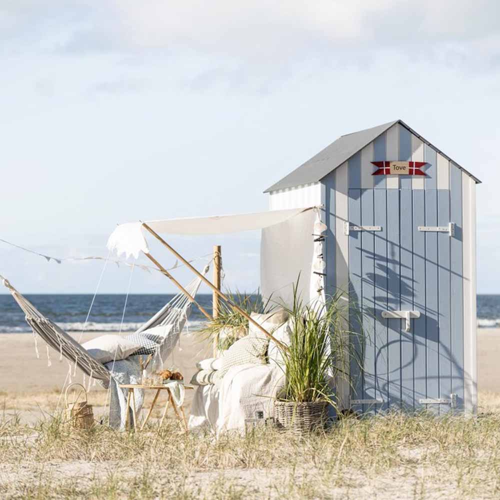 Eine Strandszene mit einer kleinen Strandhütte, einer Hängematte, Liegestühlen und Grünpflanzen auf dem Sand. Im Hintergrund ist das Meer unter einem teilweise bewölkten Himmel und einem Ib Laursen - Sonnensegel weiß mit naturfarbigen Fransen zu sehen.