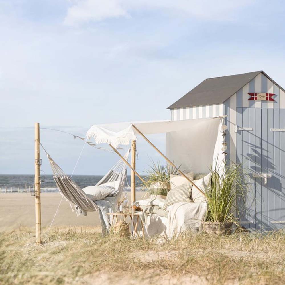 Eine kleine Strandhütte mit gestreiften Wänden steht an einem Sandstrand. Eine Hängematte, Stühle und ein Ib Laursen - Sonnensegel weiß mit naturfarbigen Fransen bilden eine gemütliche Sitzecke, geschmückt mit Pflanzen und Stranddekorationen. Im Hintergrund ist das Meer zu sehen.