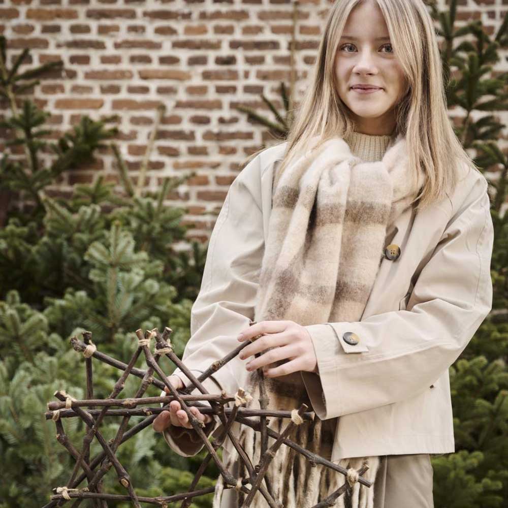 Eine Person in einem beigen Mantel und Schal steht da und hält den Ib Laursen – Stern aus Zweigen, im Hintergrund sind Kiefern und eine Backsteinmauer zu sehen.