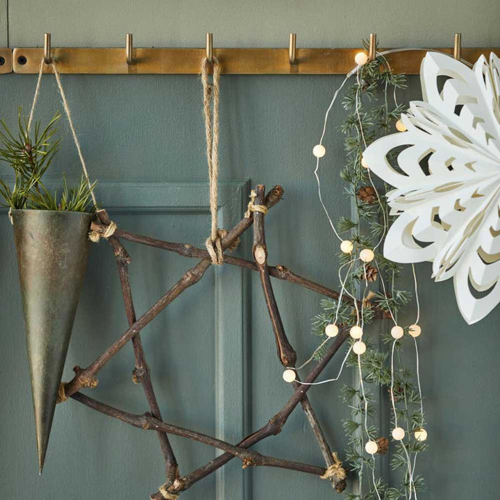 Rustikale Wanddekoration mit dem Ib Laursen – Stern aus Zweigen, aufgewertet durch eine hängende kegelförmige Metallvase, Lichterketten und eine weiße Papierschneeflocke auf grünem Hintergrund.