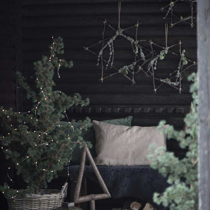 Eine gemütliche Veranda mit dem Ib Laursen – Stern aus Zweigen, einem kleinen, mit Lichtern geschmückten Tannenbaum und einer Bank mit Kissen vor einer dunklen Holzwand.