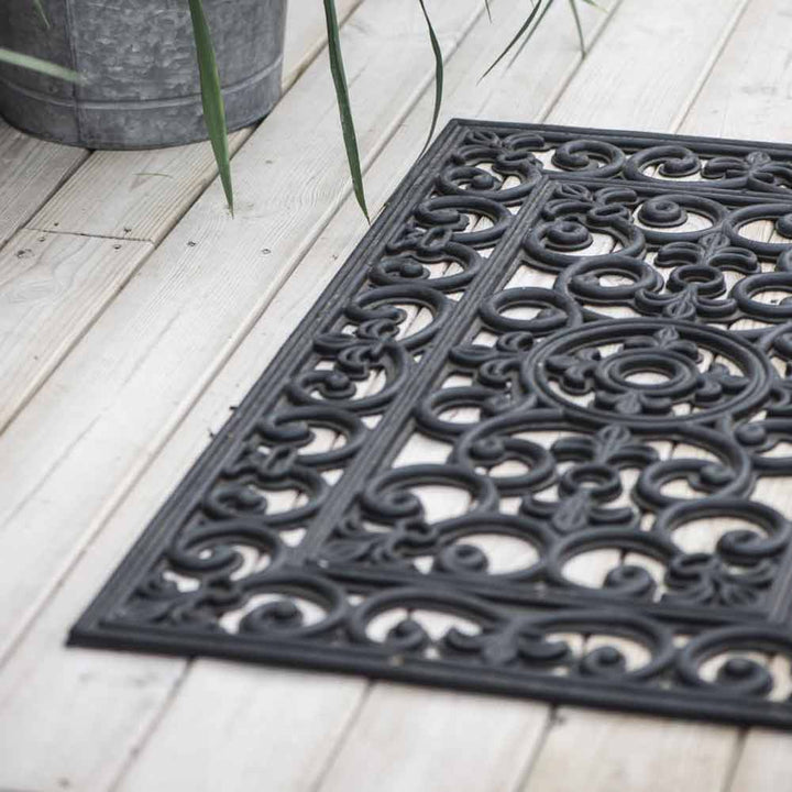 Eine elegante Ib Laursen - Türmatte Gummi Filigran mit komplizierten Mustern, platziert auf einem verwitterten Holzboden neben einem Metalleimer.