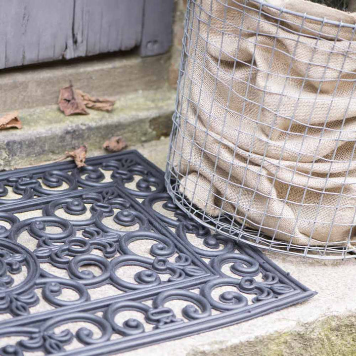 Ein Sack Kartoffeln, befestigt in einem Drahtgeflechtkäfig, neben einer Ib Laursen - Türmatte Gummi Filigran auf Betonstufen mit verstreuten Blättern.