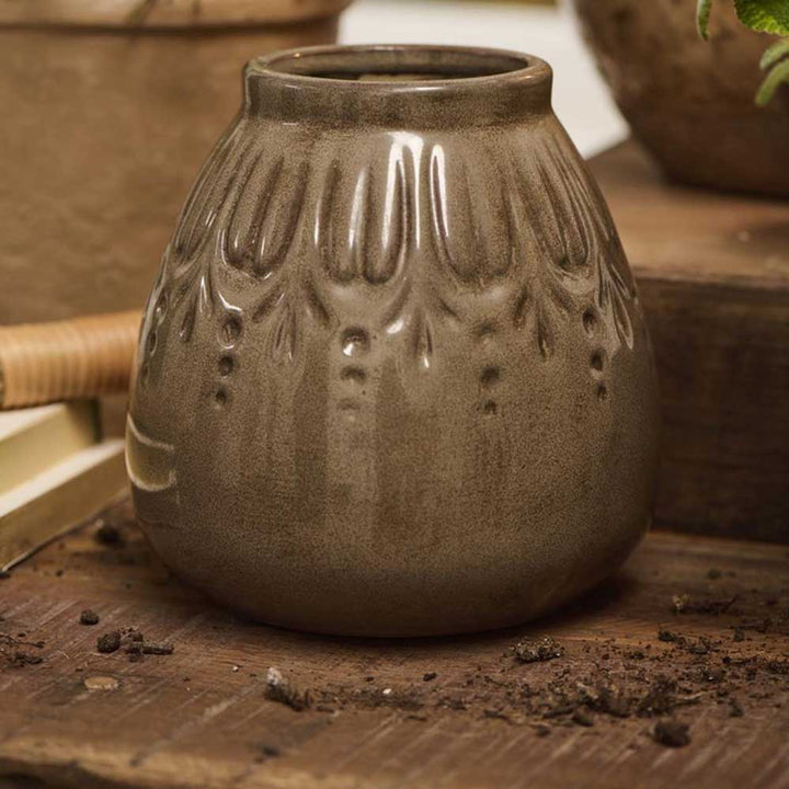 Eine kleine, braune Keramikvase von Ib Laursen mit einem strukturierten vertikalen Rillenmuster ruht auf einer Holzoberfläche, umgeben von Erde.