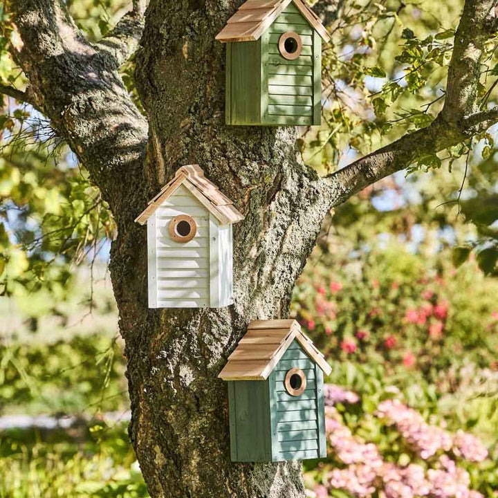 Ein Ib Laursen Vogelhaus in Weiß mit naturfarbenem Dach ist auf dem Stamm eines Baumes in einem Garten montiert.