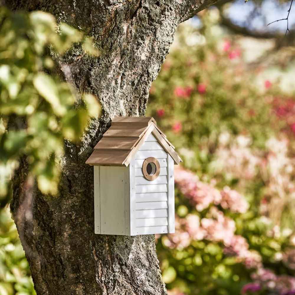 Das Ib Laursen Vogelhaus weiß mit naturfarbigem Dach, ein hölzernes Vogelhaus mit einem kleinen runden Eingang, ist auf einem Baumstamm in einem Garten mit verschwommenen Blumen im Hintergrund montiert.