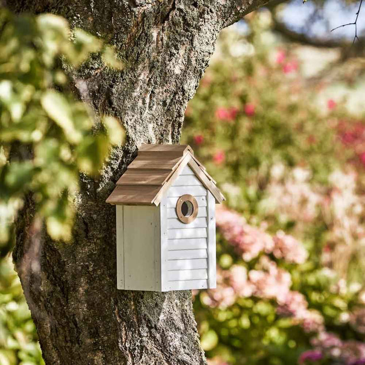 Das Ib Laursen Vogelhaus weiß mit naturfarbigem Dach, ein hölzernes Vogelhaus mit einem kleinen runden Eingang, ist auf einem Baumstamm in einem Garten mit verschwommenen Blumen im Hintergrund montiert.