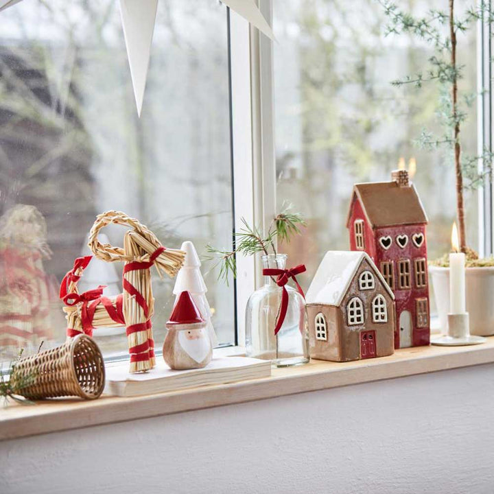 Weihnachtliches Schaufensterdekor mit der Ib Laursen - Weihnachtsziege mit rotem Band My Nostalgic, Dekofiguren, einem Lebkuchenhaus, einer Kerze und einem kleinen Ast in einer Vase.