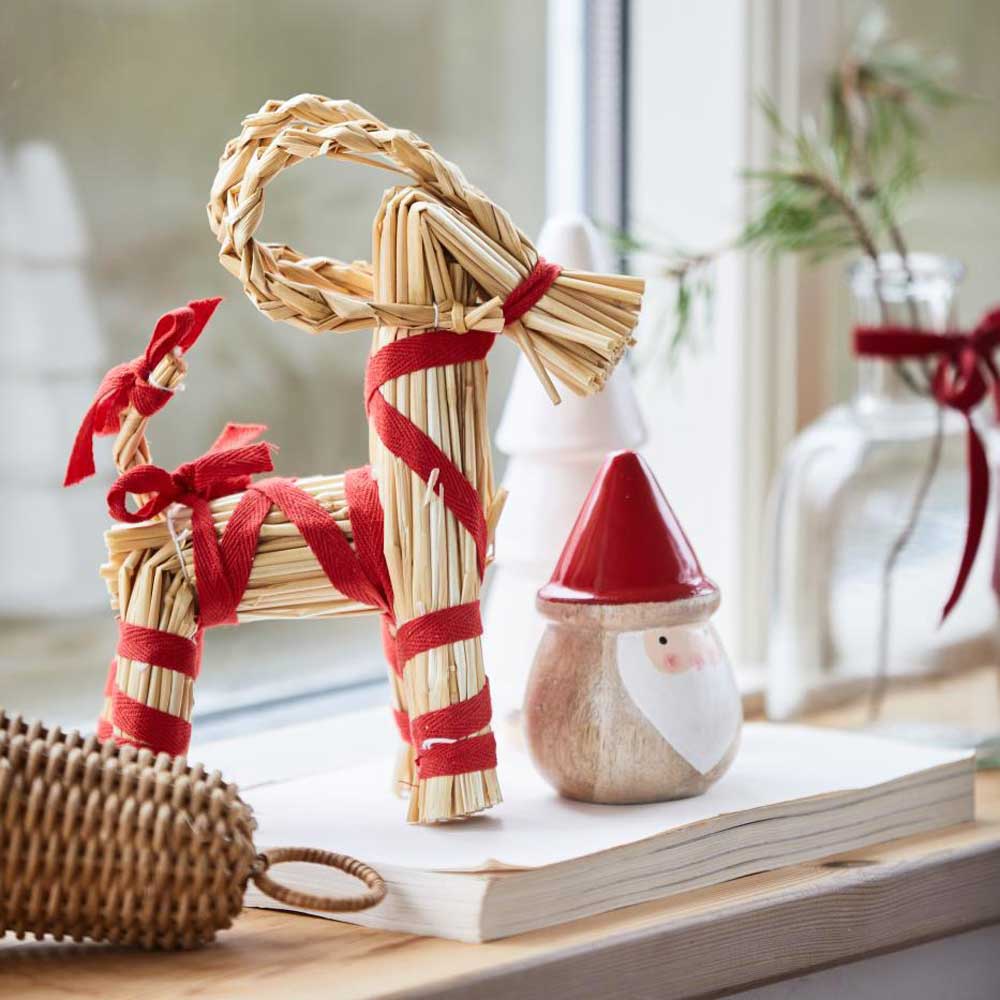 Ib Laursens Weihnachtsziege mit rotem Band „My Nostalgic“, geschmückt mit roten Bändern, sitzt neben einem kleinen Weihnachtswichtel und einer Glasflasche mit einem Tannenzweig auf einem Fensterbrett.