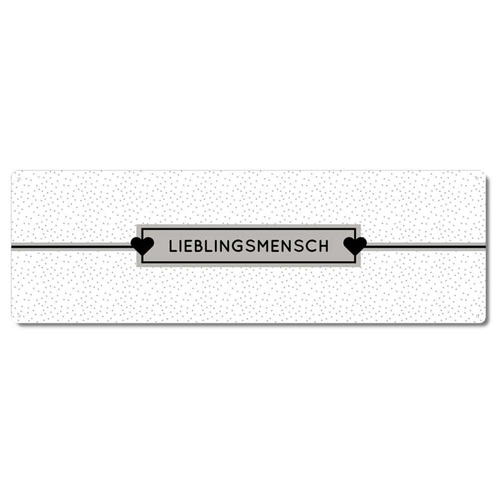 Ein rechteckiges weißes Schild mit kleinen schwarzen Punkten und der Aufschrift „Interluxe Metallschild – Lieblingsmensch“ in der Mitte, flankiert von zwei schwarzen Herzen.