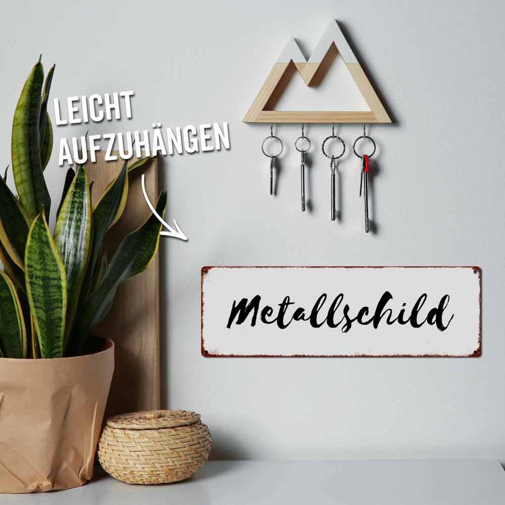 An einer Wand über einer Pflanze und einem Korb hängt ein Schlüsselbrett mit vier Schlüsseln. Auf einem Schild daneben steht „Interluxe Metallschild mit Flaschenöffner – Universalschlüssel nach DIN 0815“. Der Text „Leicht aufzuhängen“ weist auf das Schlüsselbrett hin.