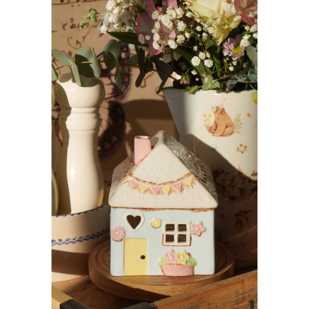 Das Isabelle Rose - Keramik Haus als Teelichthalter im Landhausstil Pale Blue steht auf einer Holzoberfläche, umgeben von Blumen und einer weißen Vase mit Bärenmotiv.