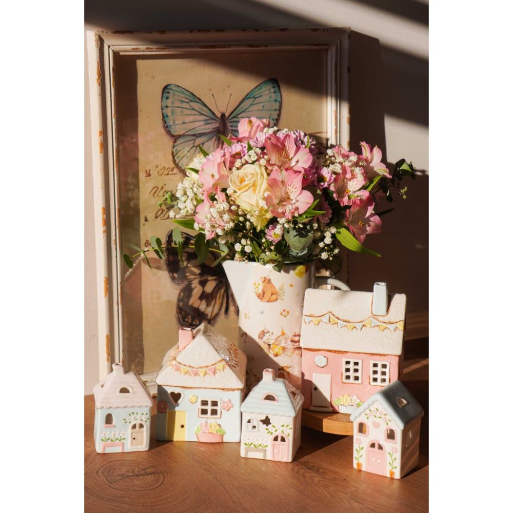 Ein Isabelle Rose - Keramik Haus als Teelichthalter im Landhausstil Rosa steht auf einer hölzernen Unterlage, die mit einem Blumenkrug und einem Schmetterling bedruckt ist. Vier dieser kleinen Keramik-Haus-Teelichthalter umgeben das Arrangement.